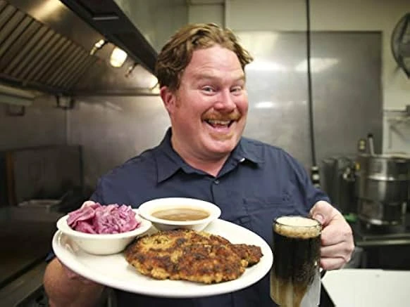 A grinning red-haired man with a mustache holds a plate of breaded cutlet, red cabbage and gravy and a frothy dark drink in a kitchen.