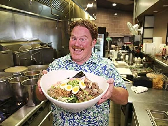 A grinning red-haired man with a mustache in a blue print shirt holds out a large bowl topped with halved eggs, meat and greens.