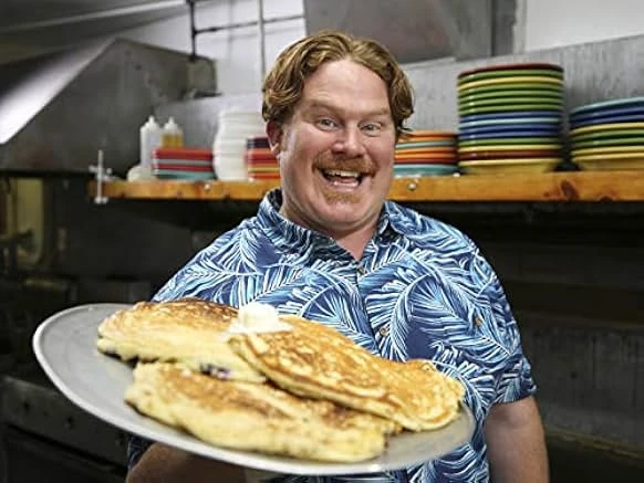 A grinning red-haired man with a mustache in a blue palm-print shirt holds a platter of golden pancakes in a restaurant kitchen.