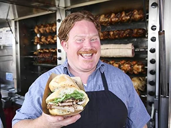 A grinning red-haired man with a mustache in a denim shirt and black apron holds a sandwich in front of racks of rotisserie chickens.