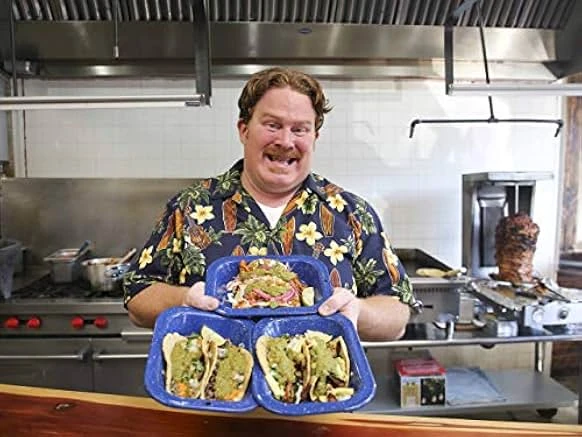 A red-haired man with a mustache in a floral shirt holds three blue enamel plates of tacos with green salsa in a restaurant kitchen.