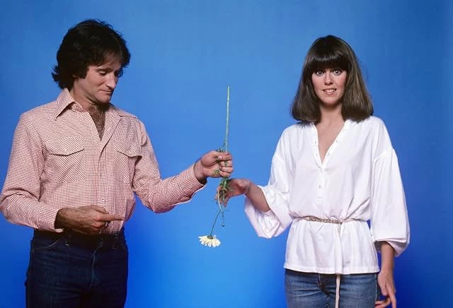 A man in a pink checked shirt hands a wilting daisy to a smiling woman in a white blouse against a bright blue backdrop