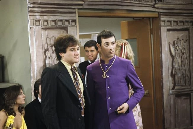 A curly-haired man in a dark suit and patterned tie stands beside a man in a purple tunic and peace-sign medallion in a doorway