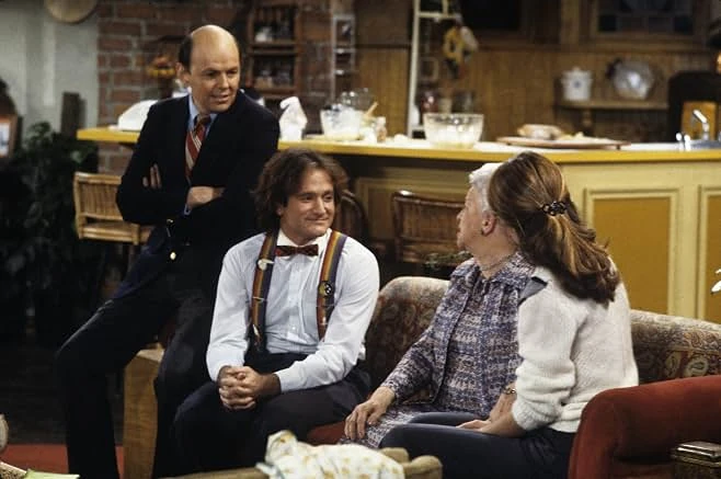 Mork in rainbow suspenders and a bow tie sits on a sofa with two women as a balding man in a blazer stands behind him