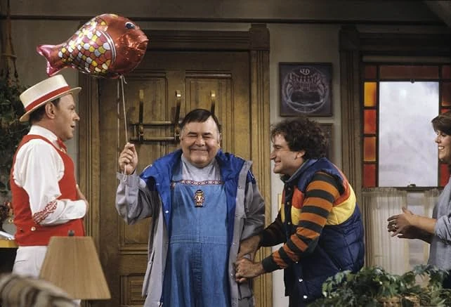 Mork in a striped sweater and vest laughs with a man in overalls holding a fish-shaped balloon as a man in a straw boater watches