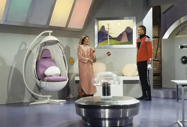 A man in a striped nightshirt and cap stands in a futuristic room with an egg chair as a man in a red uniform looks on