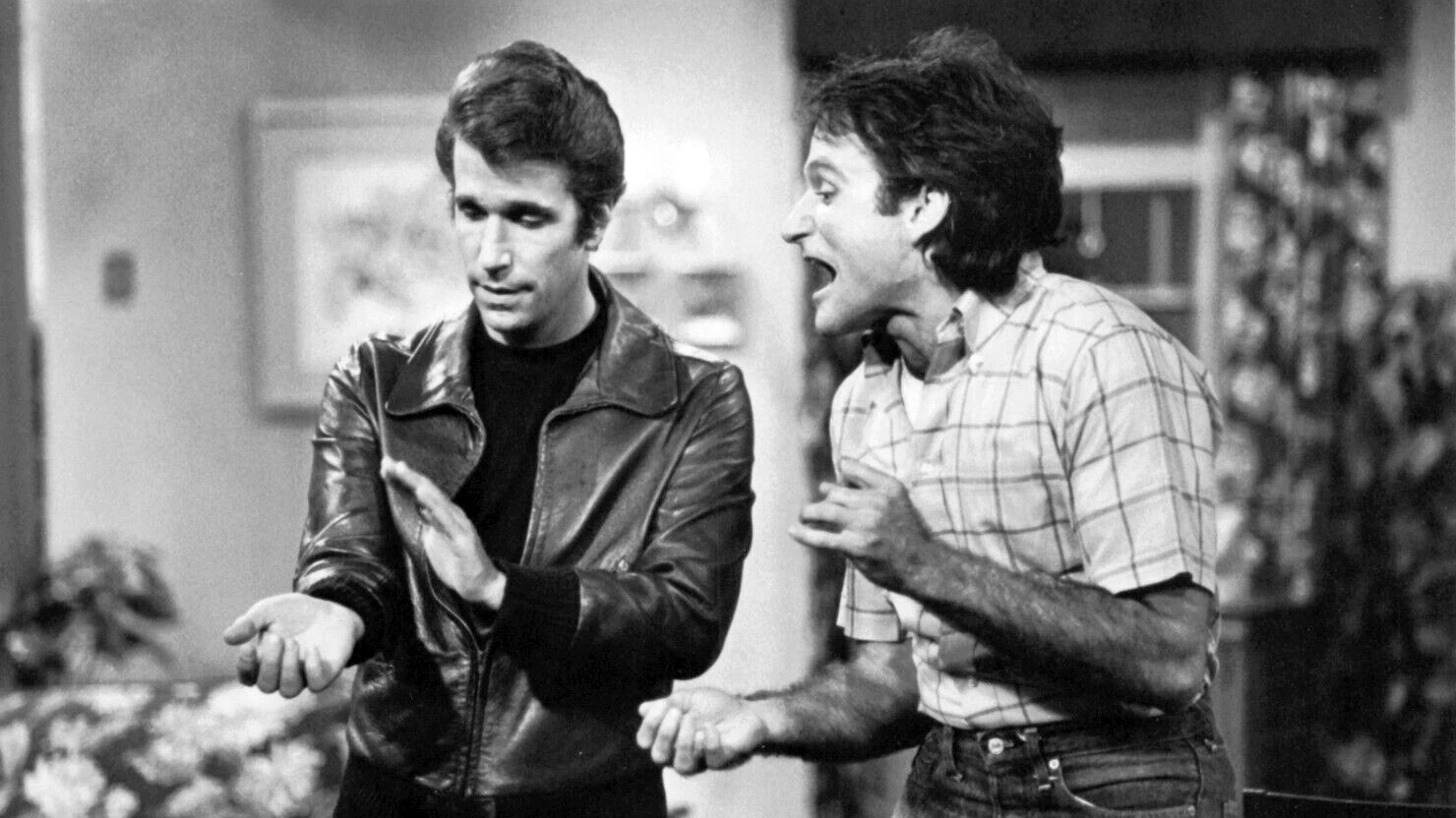 Robin Williams as Mork shouting beside Henry Winkler as Fonzie in a leather jacket on a living room set, black and white
