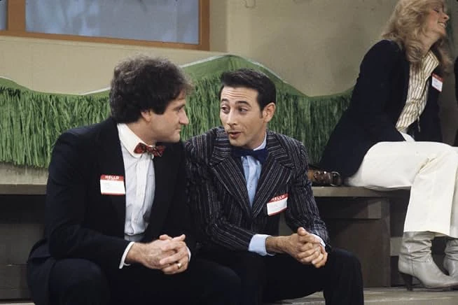 Two men in dark suits, bow ties and HELLO name tags sit talking on a low ledge as a woman in a blazer and white trousers sits nearby