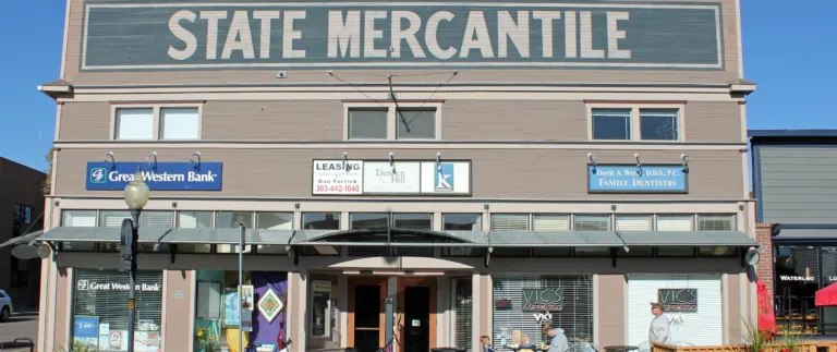 Two-story tan wood-frame building with State Mercantile painted in large cream letters across its false front, under a bracketed cornice.