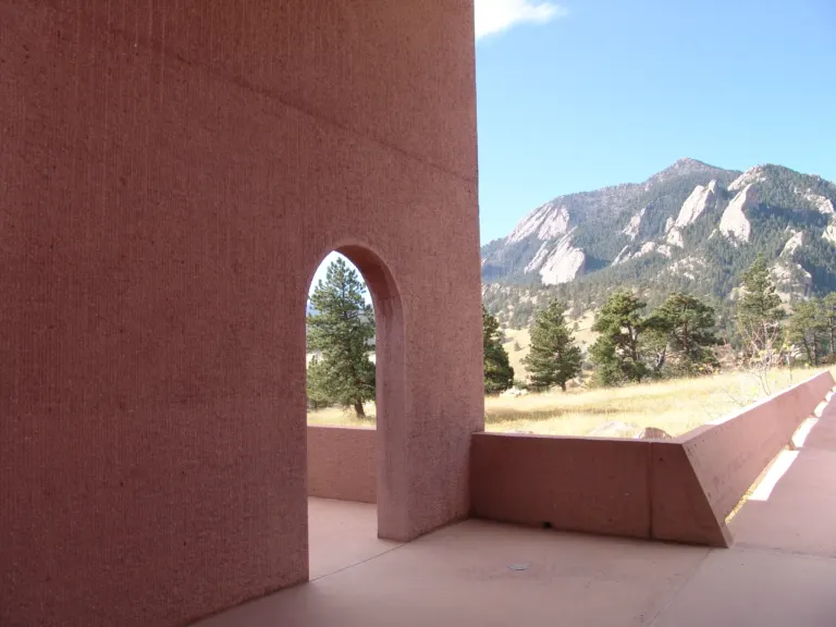Curved pink concrete wall with an arched opening on a terrace at the NCAR Mesa Laboratory, facing pine trees and a rocky peak