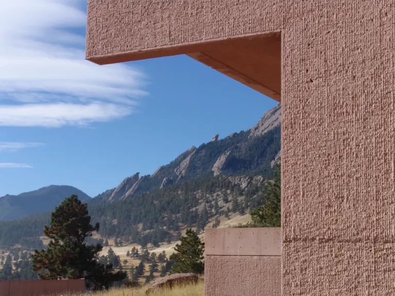 Rough-textured pink concrete corner of the NCAR Mesa Laboratory framing the Flatirons, pine trees and streaked clouds