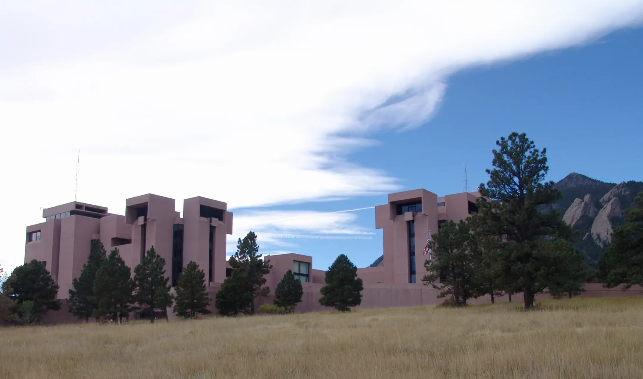 Pink concrete towers of the NCAR Mesa Laboratory behind pine trees and a golden grass meadow, with the Flatirons at right