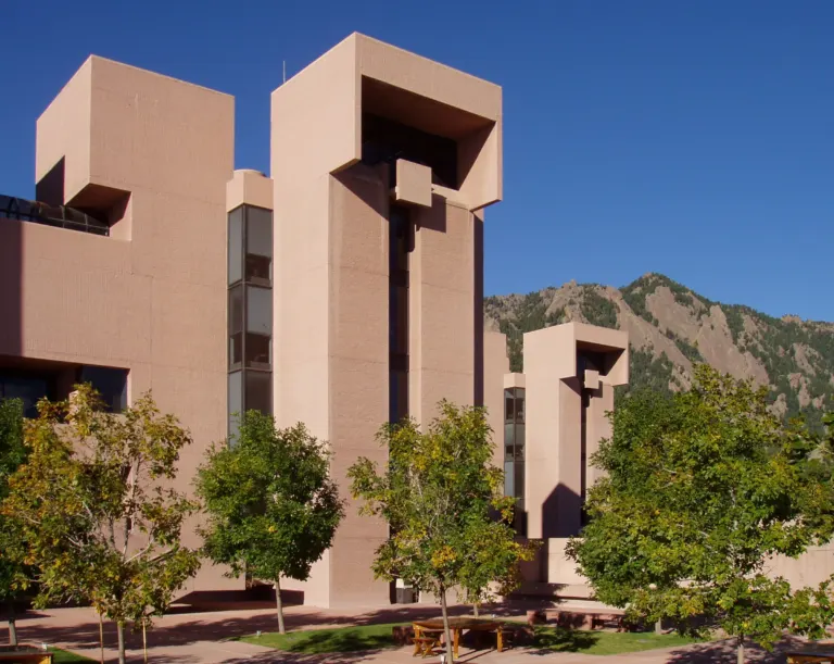 Hooded pink concrete towers of the NCAR Mesa Laboratory against a deep blue sky, with a Flatirons peak behind and trees below