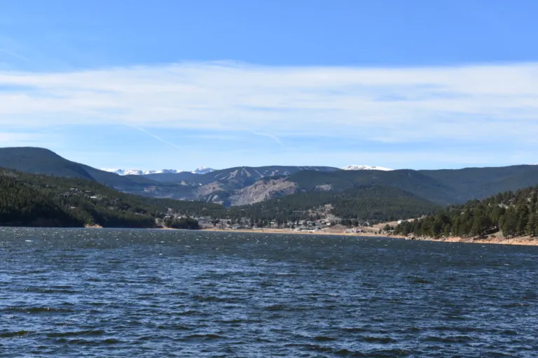 Wind-ruffled reservoir with a small town on the far shore below forested hills, ski runs and distant snow-capped peaks.