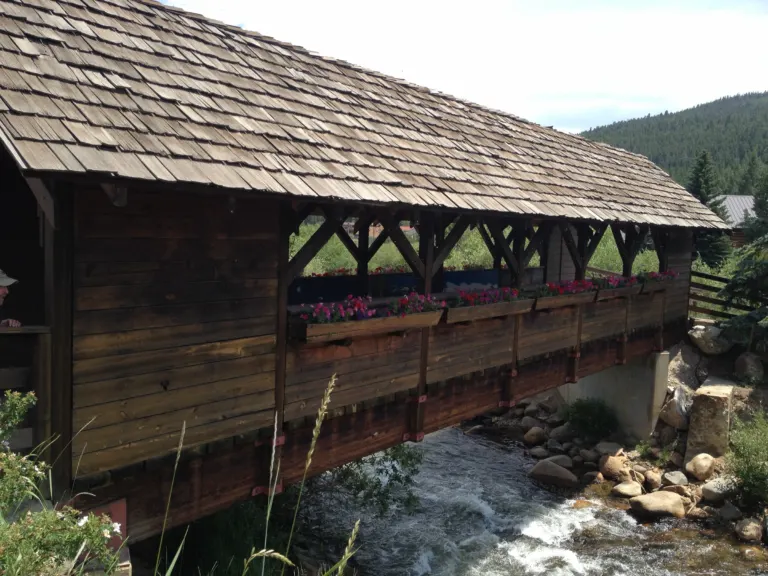 Wooden covered footbridge with a shake roof and flower boxes spanning a rocky creek, with a forested hill behind.