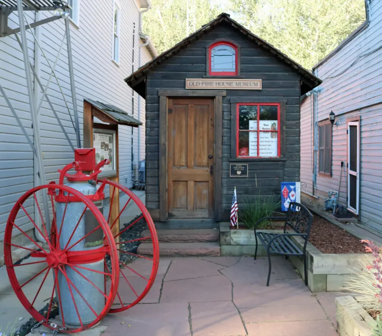 Small dark wooden firehouse with red window trim between two clapboard buildings, with a red-wheeled antique fire cart in front.