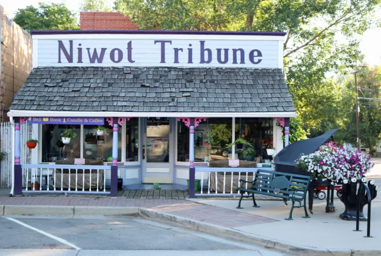 White wooden storefront with a shingled porch roof and Niwot Tribune in purple letters, beside a piano planter full of petunias.