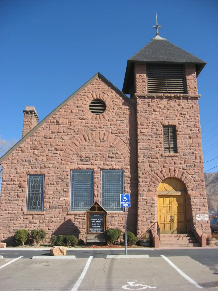 Red sandstone church with a square bell tower and arched wooden doors under a clear blue sky, seen across a small parking lot.