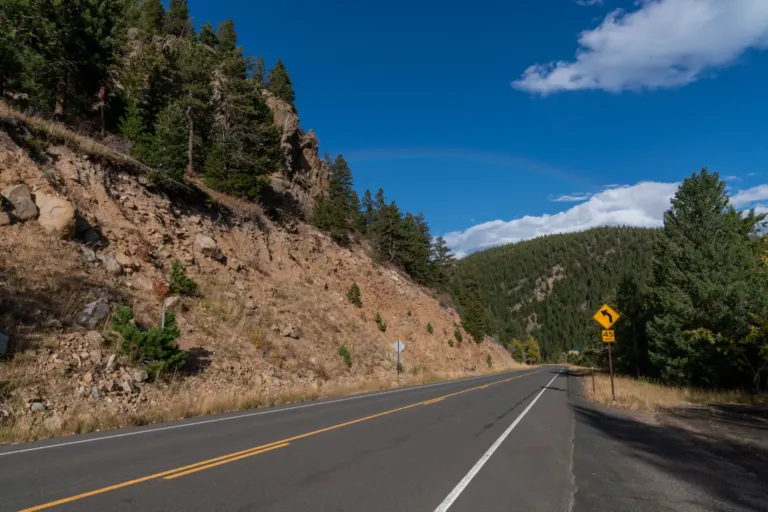 Empty two-lane mountain highway beside a rocky road cut topped with pines, with a yellow curve warning sign and a forested hill ahead