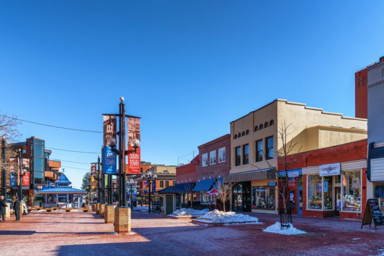 Brick pedestrian mall with banner poles, a blue-roofed kiosk and low storefronts under a clear blue sky, with patches of snow.