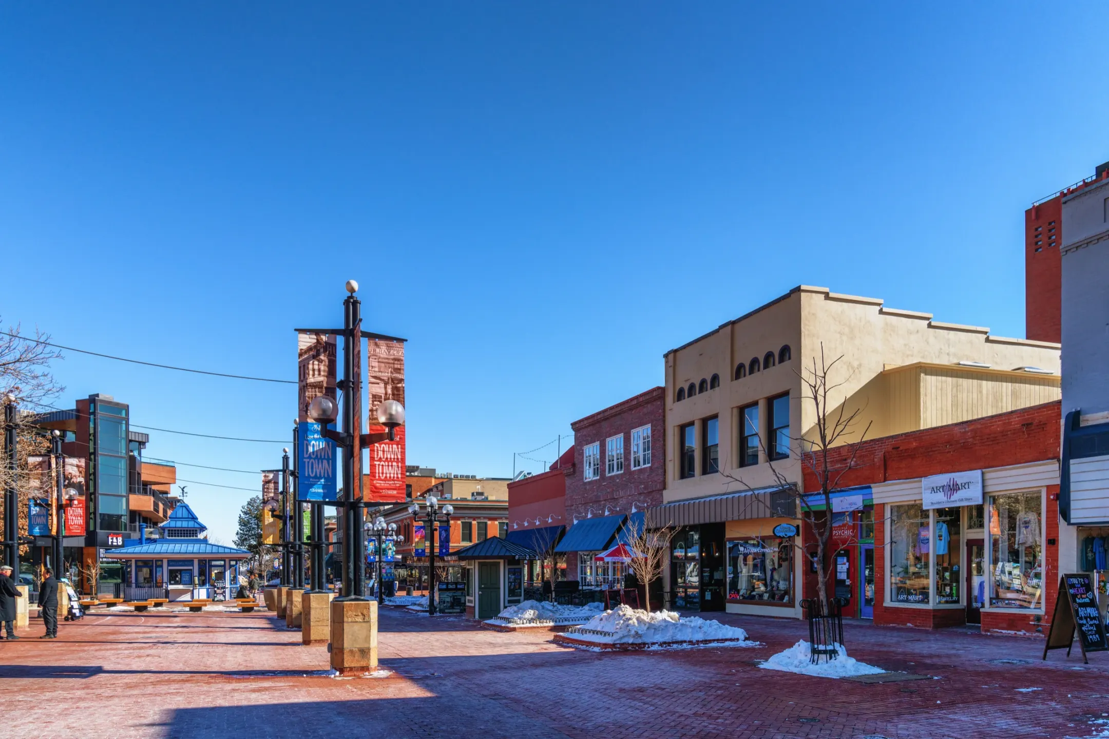 Brick pedestrian mall with banner poles, a blue-roofed kiosk and low storefronts under a clear blue sky, with patches of snow.