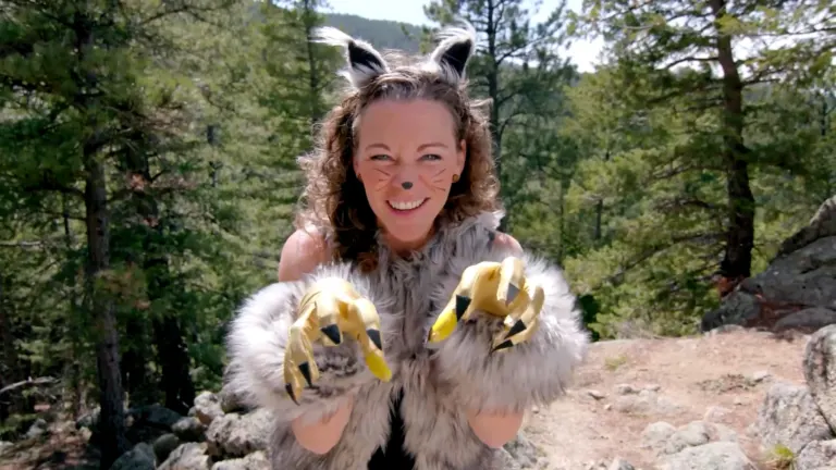 A smiling woman in a bobcat costume with fur ears, whisker face paint and golden claw gloves reaches toward the camera in a pine forest
