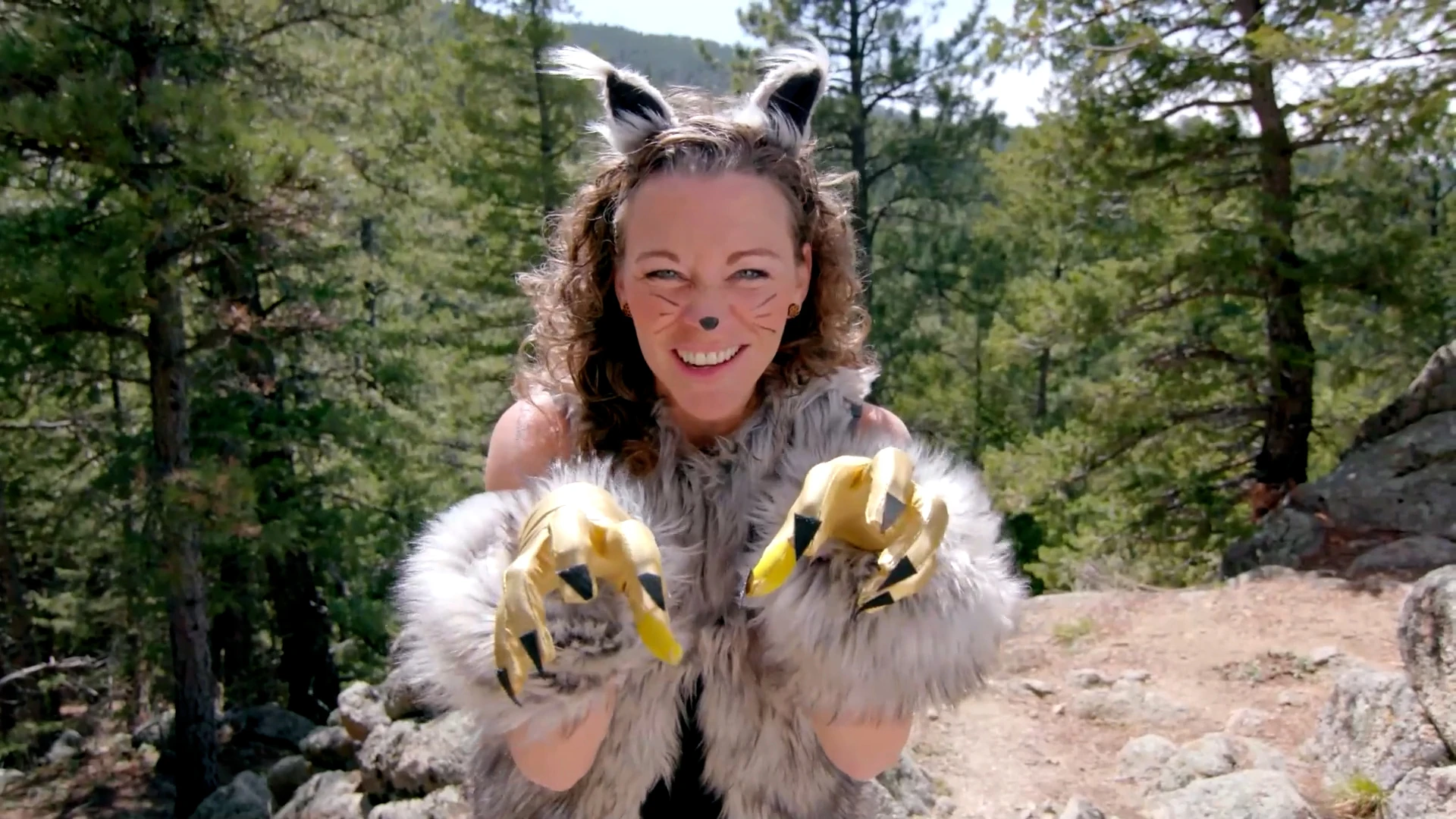 A smiling woman in a bobcat costume with fur ears, whisker face paint and golden claw gloves reaches toward the camera in a pine forest