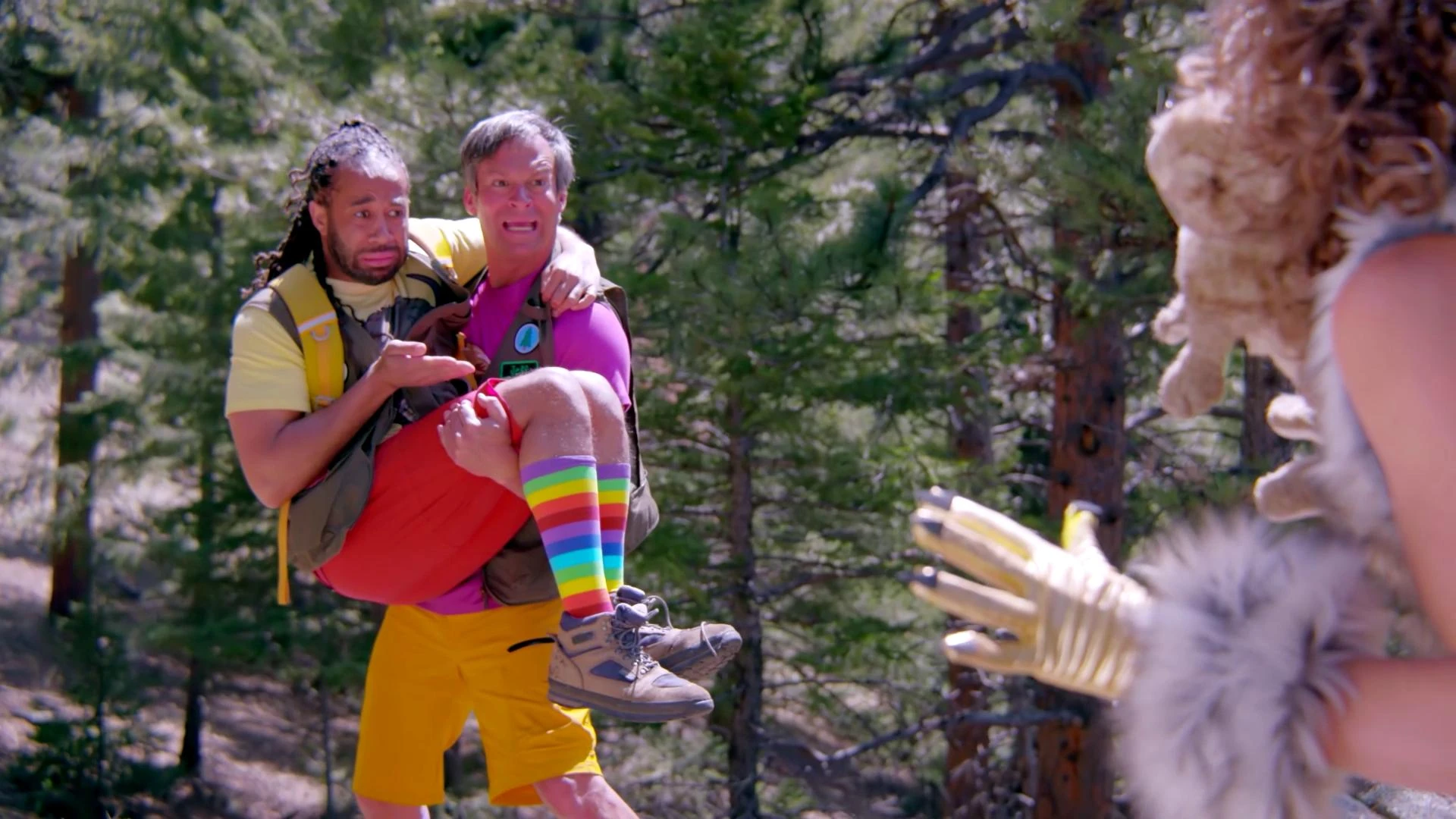 A gray-haired man in a magenta shirt carries a man in rainbow socks through a pine forest as a bobcat claw reaches in from the right