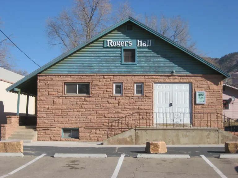 Gabled hall with red sandstone walls, a green wooden gable lettered Rogers Hall and white double doors, facing an empty parking lot.