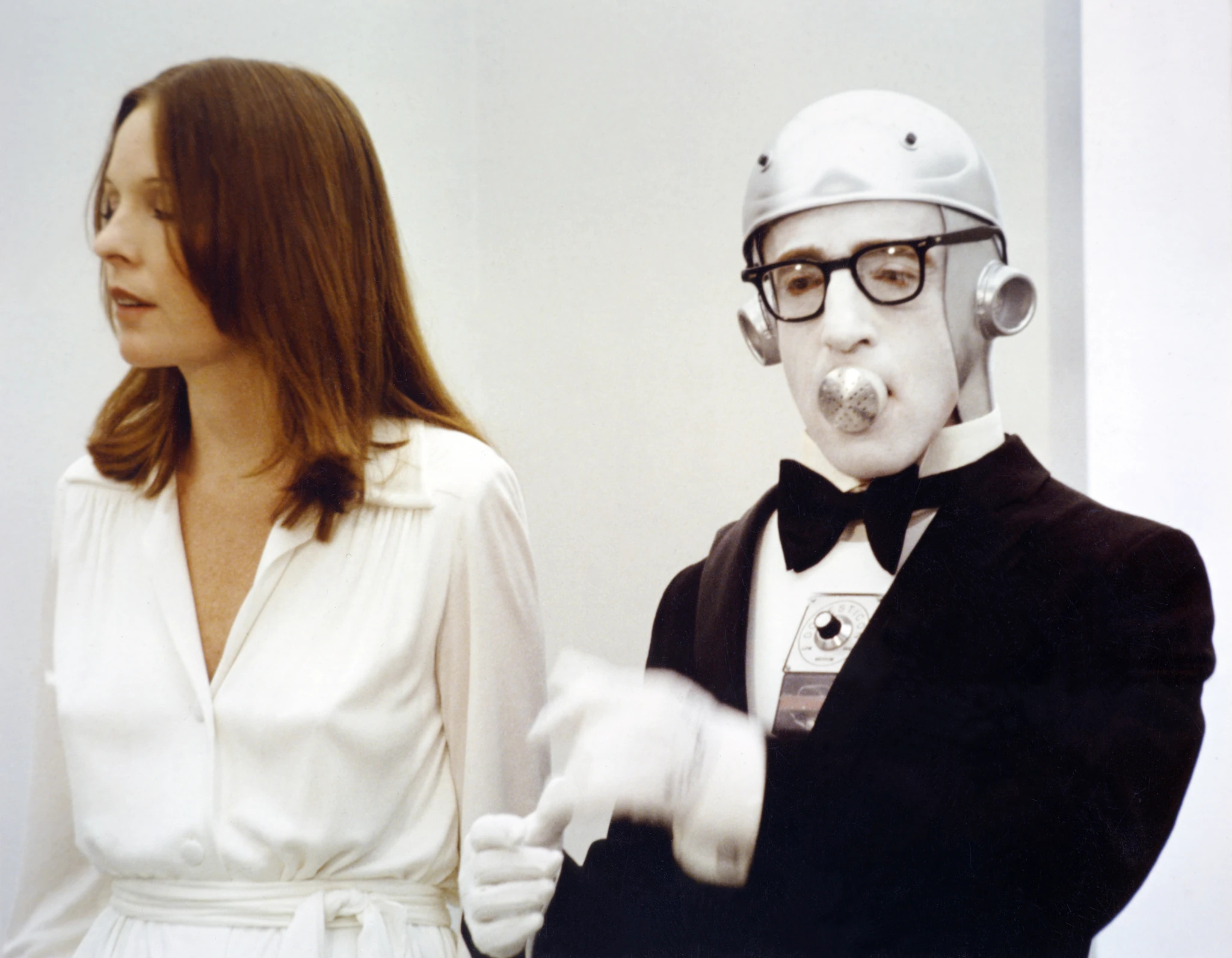 Woody Allen as Miles, disguised as a robot butler in a silver helmet, glasses and bow tie, beside a woman in a white dress