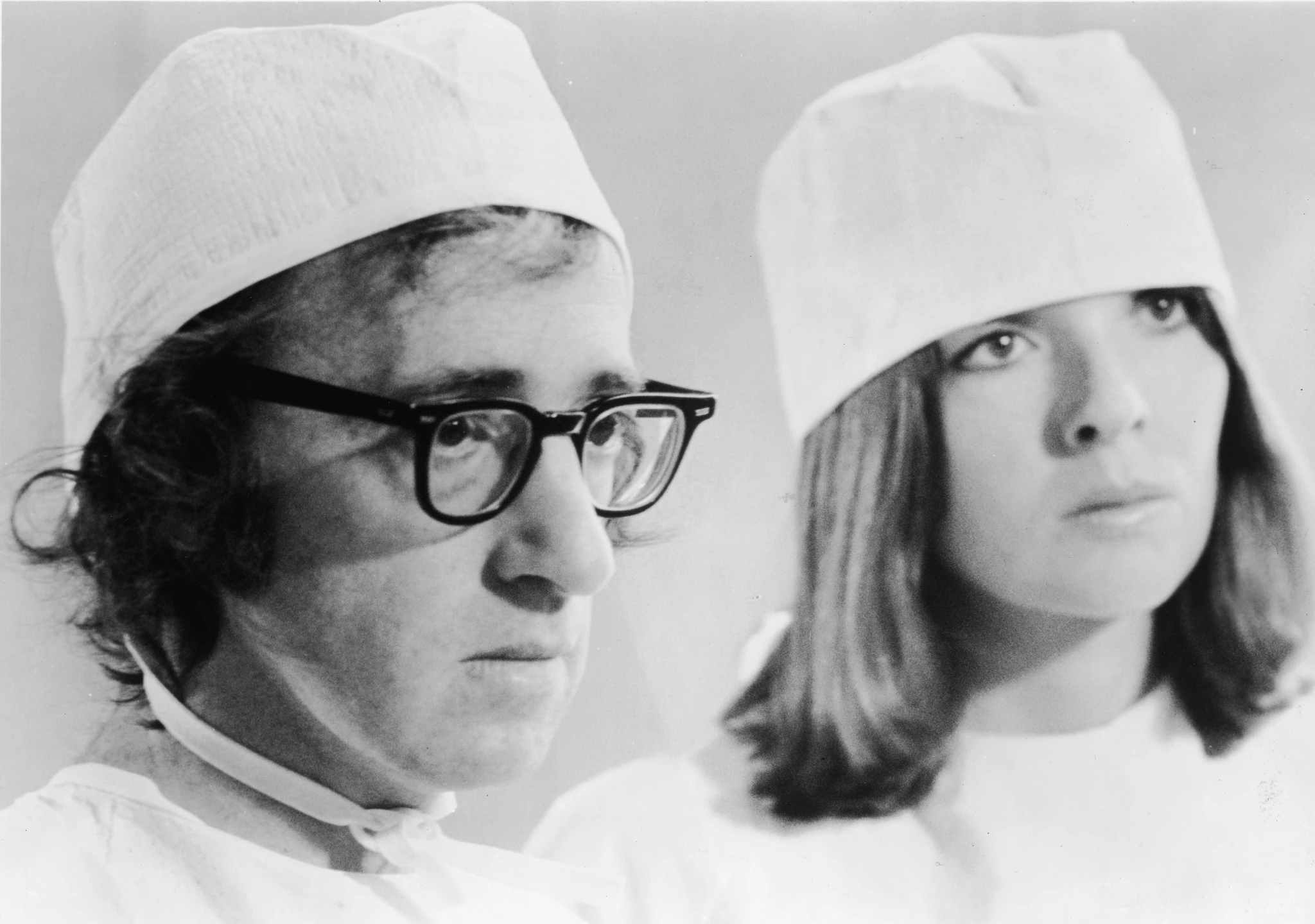 Close-up of a man in heavy black glasses and a woman with straight hair, both in white surgical caps, gazing to the left