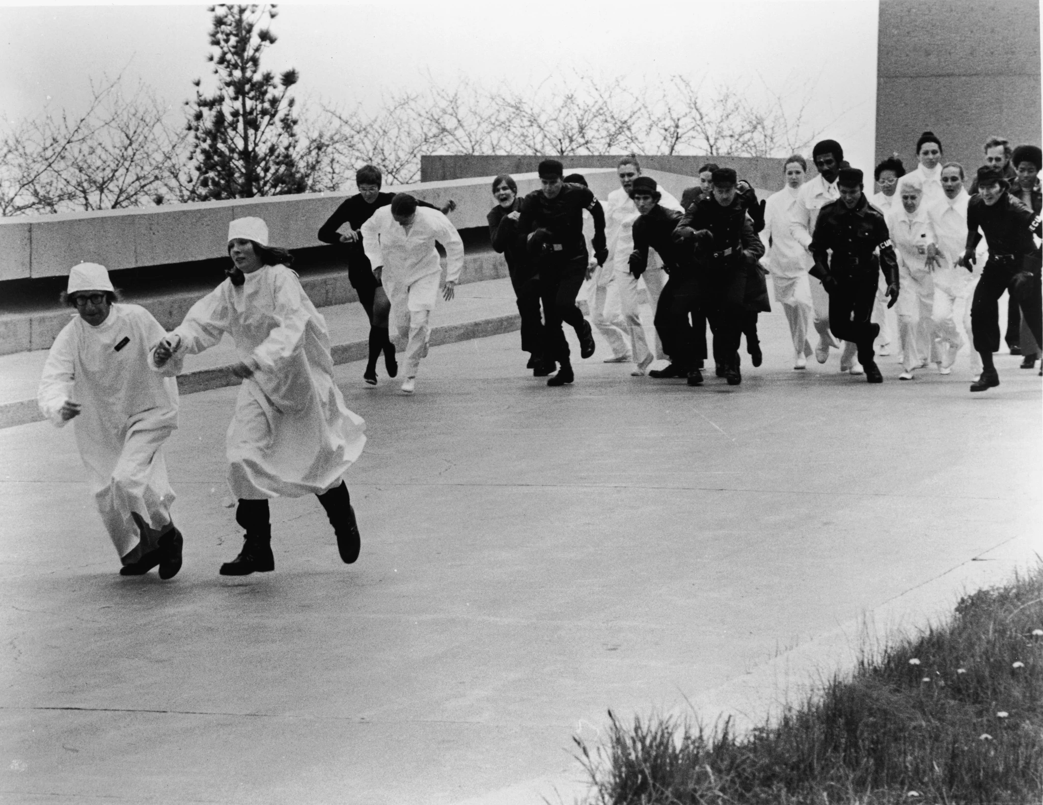 Two people in white surgical gowns and caps run across a concrete plaza, chased by guards in black and staff in white