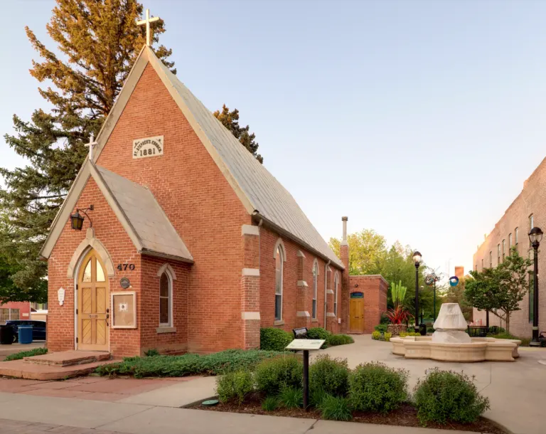 Small red brick church with a steep roof and a pointed-arch wooden door beside a paved plaza with a covered fountain, in warm evening light.