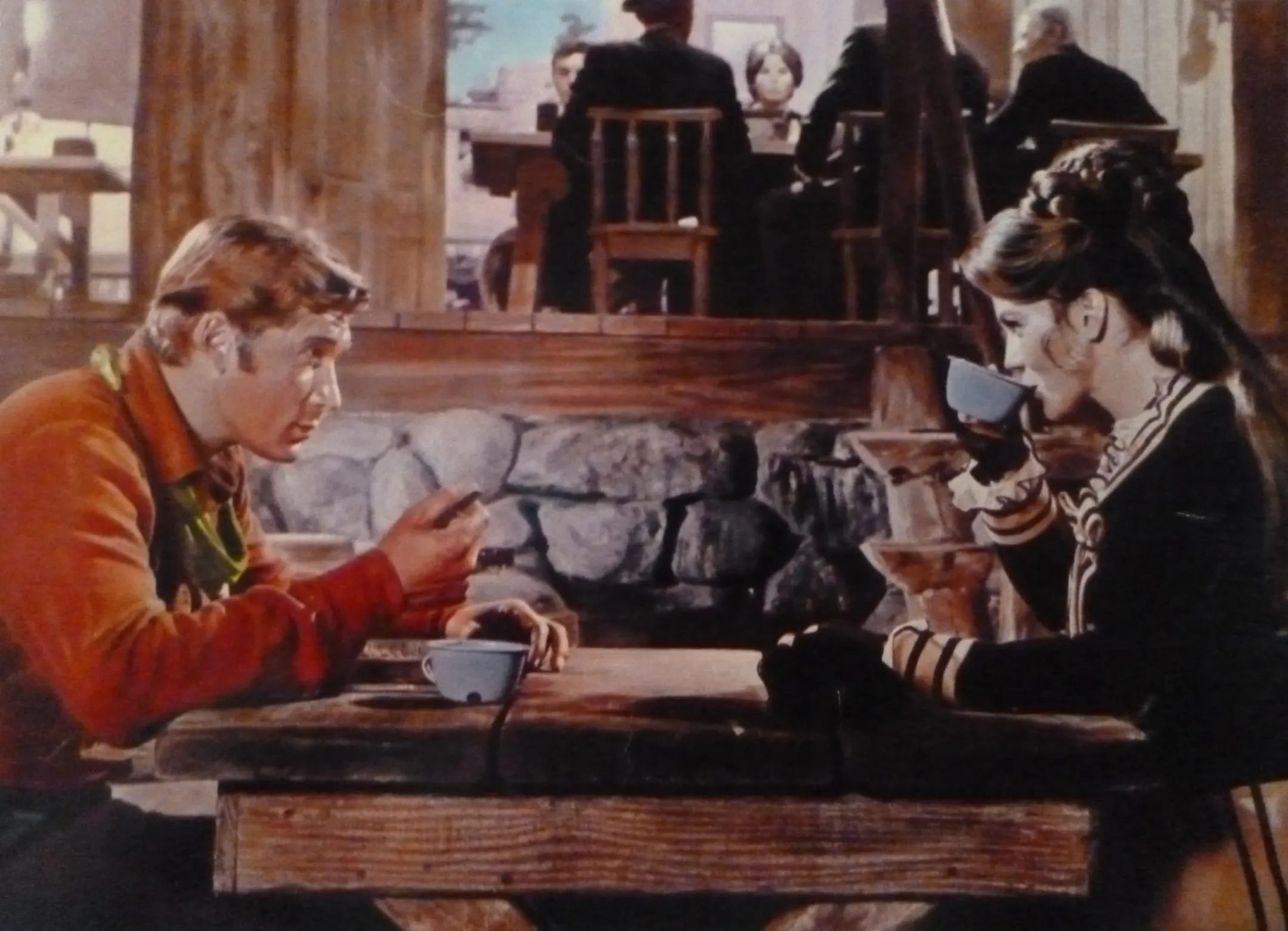 A man in an orange shirt talks across a rough wooden table to a woman in a dark jacket sipping from a tin cup.