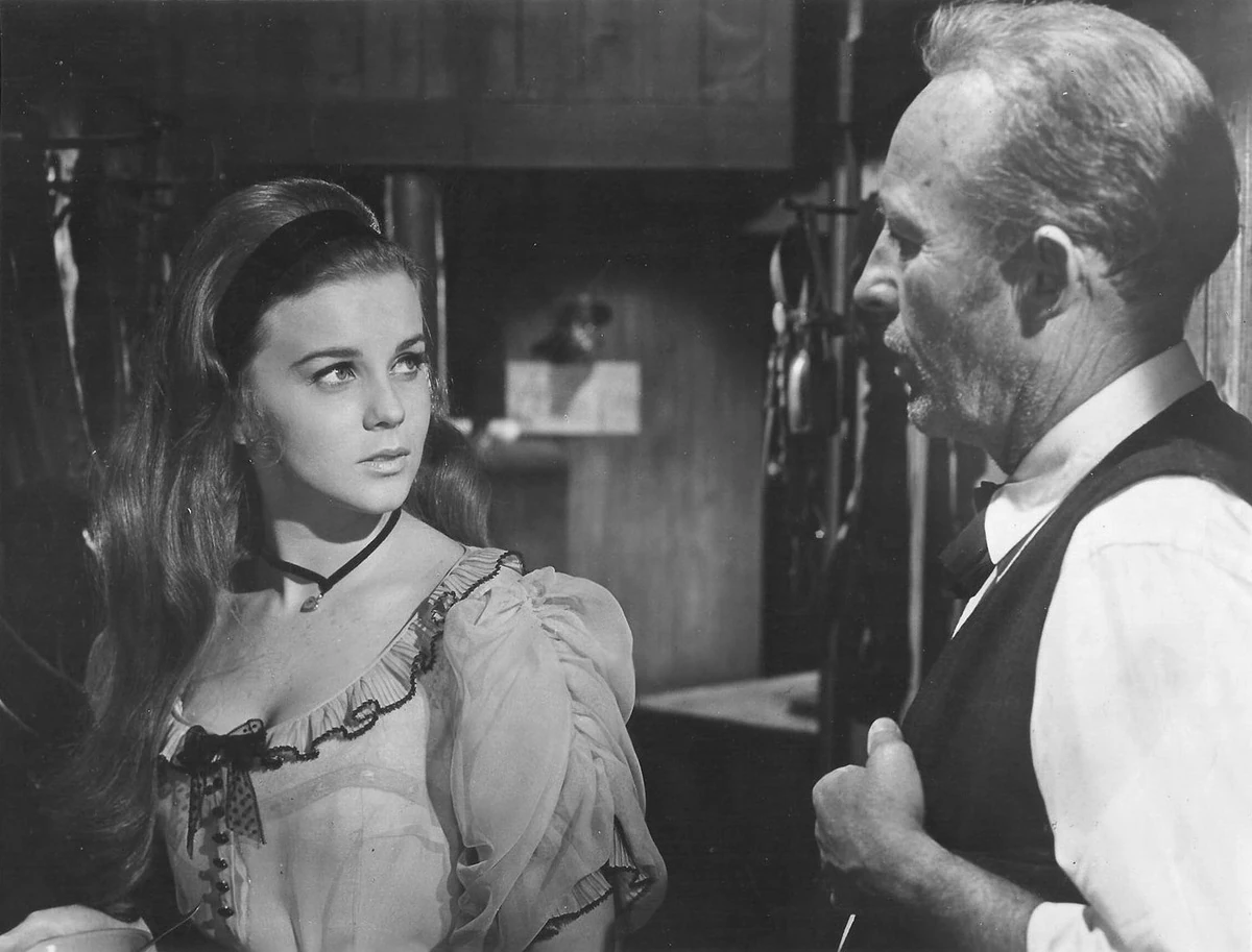 A young woman with long hair and a velvet choker looks up at a gray-haired man in a vest and bow tie in a wooden room.