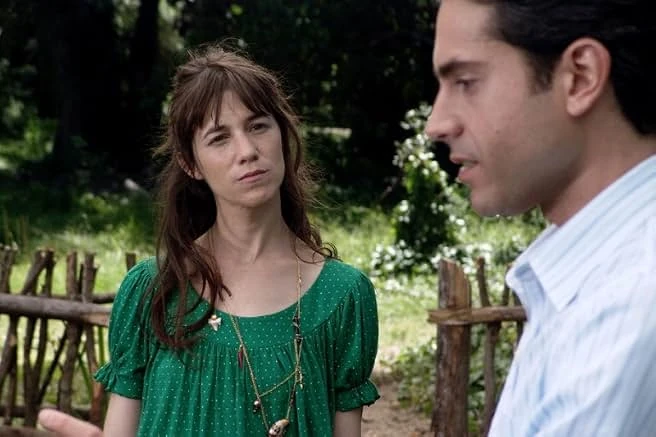 A woman in a green polka-dot dress listens to a man in a striped shirt beside a rustic wooden fence in a garden