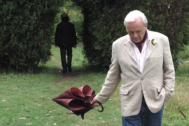 A white-haired man in a cream linen jacket holds a red umbrella on a lawn as a dark figure walks away between hedges