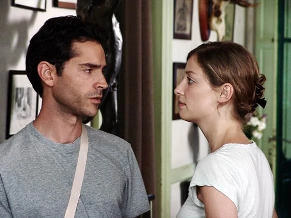 A man in a gray T-shirt and a woman in a white top face each other in a hallway hung with framed pictures