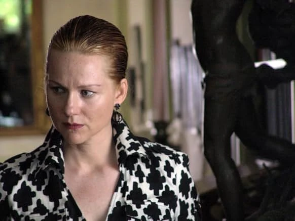 A woman with slicked-back red hair in a black-and-white patterned blouse glances sideways beside a dark bronze statue