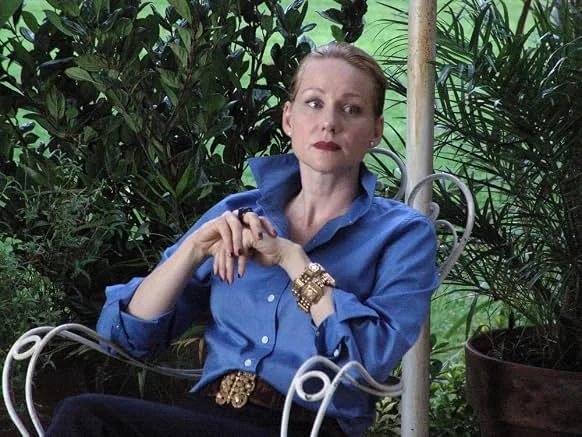 A woman in a blue shirt and a jeweled gold cuff bracelet sits on a white wrought-iron chair among potted palms