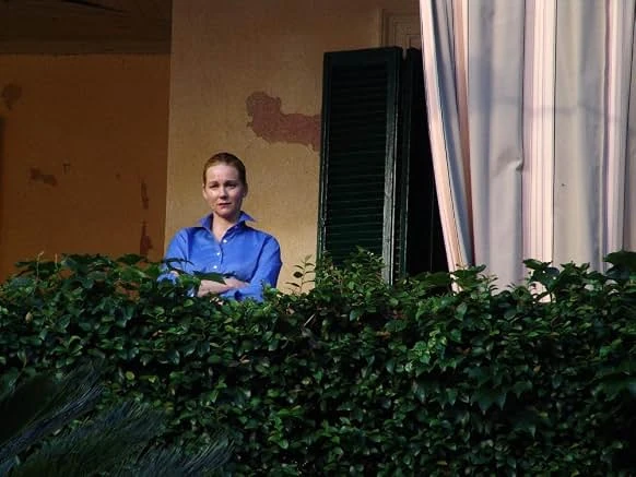 A woman in a blue shirt stands with arms crossed on a balcony behind a tall hedge, beside green shutters and a curtain