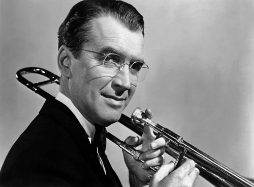 James Stewart as Glenn Miller, in rimless glasses and a dark suit, smiles with a trombone resting on his shoulder.
