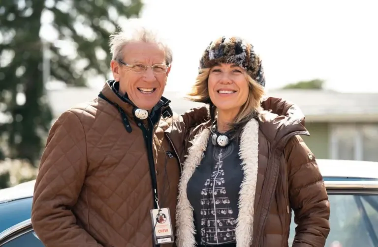 A smiling white-haired man with glasses and a crew badge stands with a woman in a fur hat and brown parka beside a vintage car