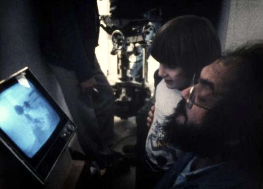 A bearded man in glasses and a young boy watch a small video monitor on a dim film set, a camera rig behind them