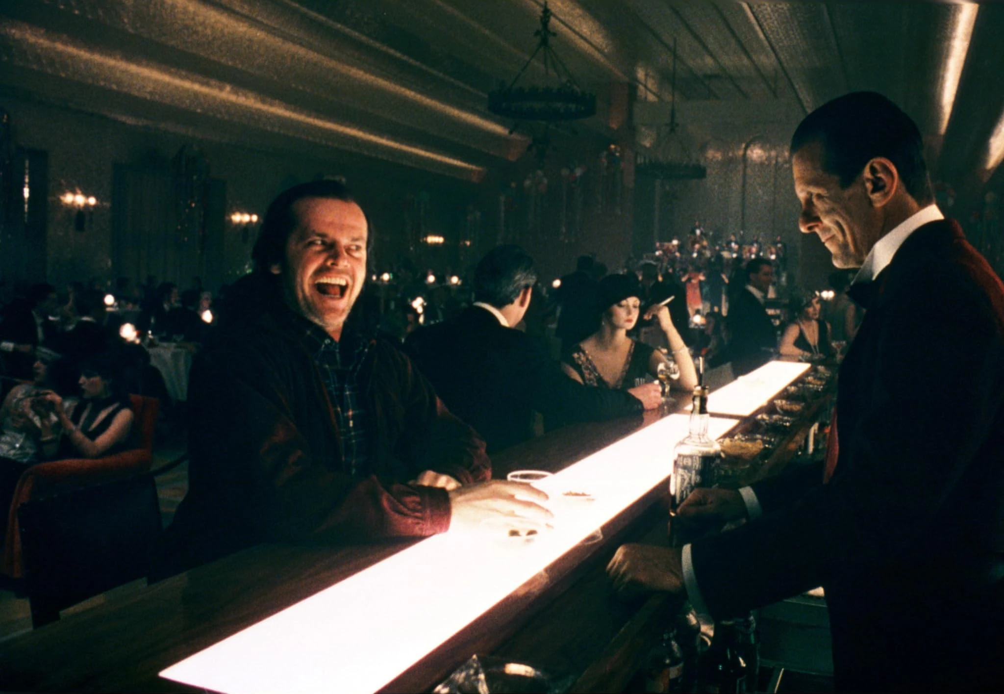 Jack Torrance laughs at the glowing Gold Room bar as a bartender in a red jacket serves him, with 1920s partygoers behind