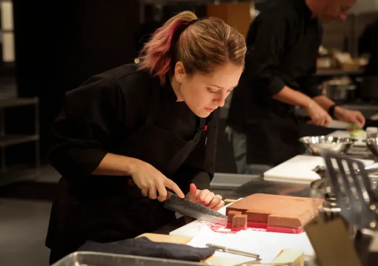 A chef with a pink-streaked ponytail in a black chef's jacket carefully slices a layered pink and brown dish in a busy kitchen.