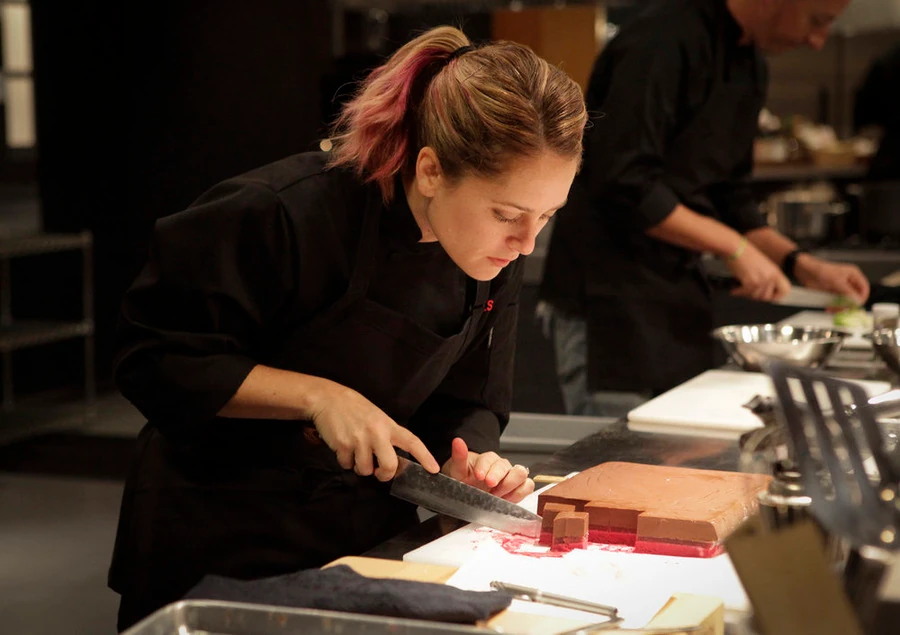 A chef with a pink-streaked ponytail in a black chef's jacket carefully slices a layered pink and brown dish in a busy kitchen.