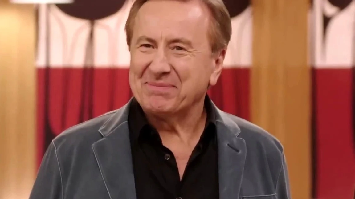 A smiling man with graying light brown hair in a gray velvet jacket and black shirt stands in front of a red and white backdrop.
