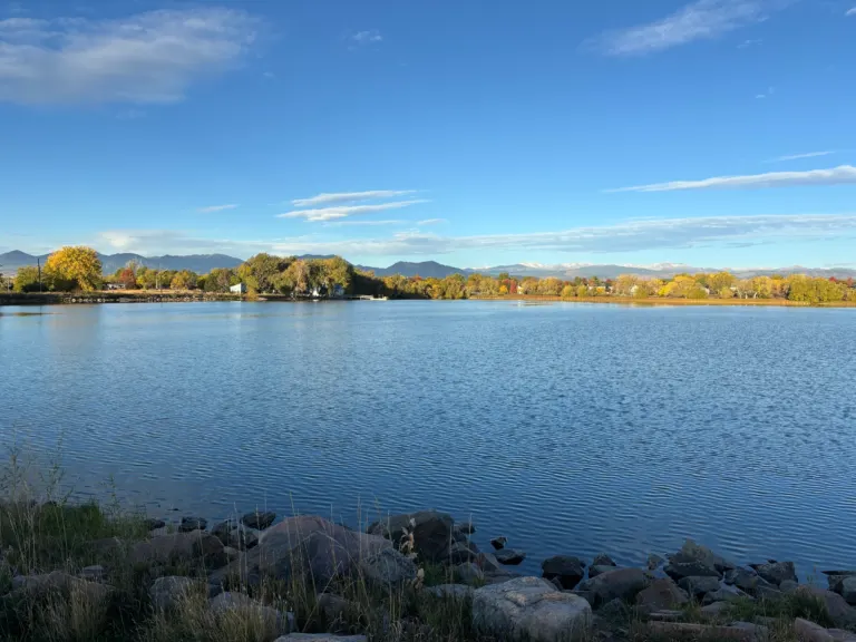 Blue lake with shoreline rocks in the foreground, autumn cottonwoods on the far shore and snow-dusted mountains under a blue sky.