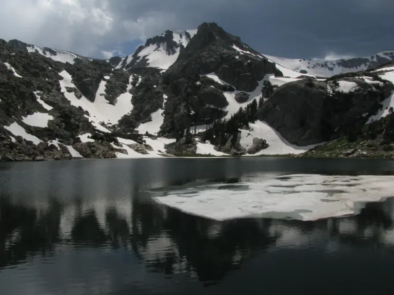 A dark alpine lake with a floating sheet of ice below a rocky, snow-patched peak under storm clouds.